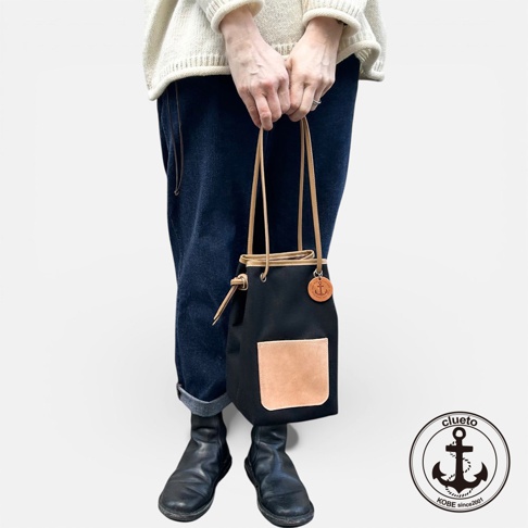 一個穿著米白色毛衣和深藍色牛仔褲的人，左手拿著一個黑色束口包，包上有一個皮革口袋和一個圓形吊牌。