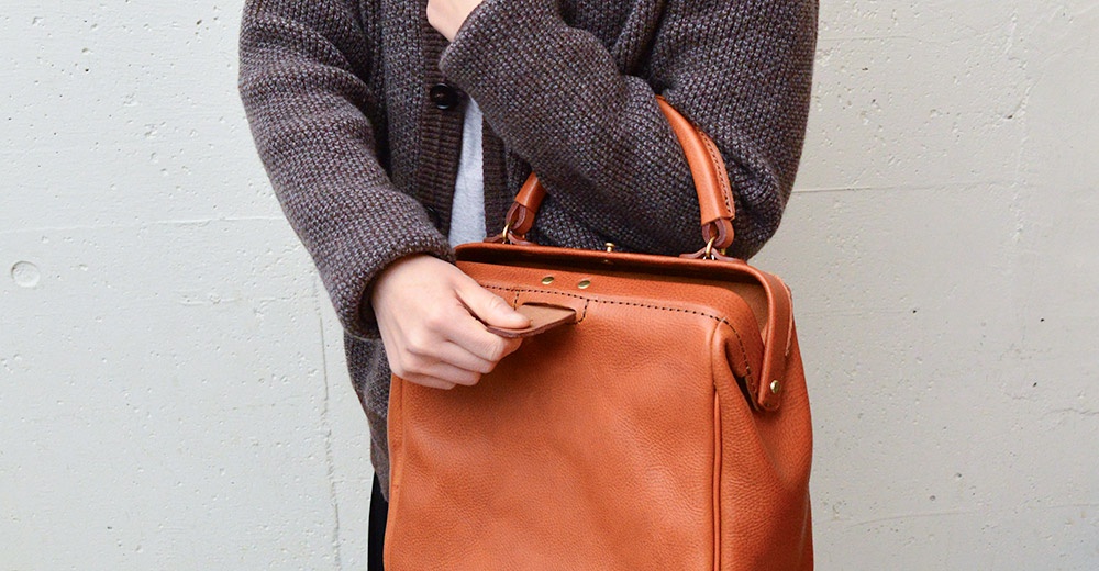 棕色皮革製的方形口金托特包，包身有縫線細節，上方有提把，搭配灰色紋理外套。