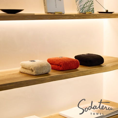 養育TOWEL 三色浴巾，以簡約的風格陳列在木質層架上，圖片下方有品牌 Logo。