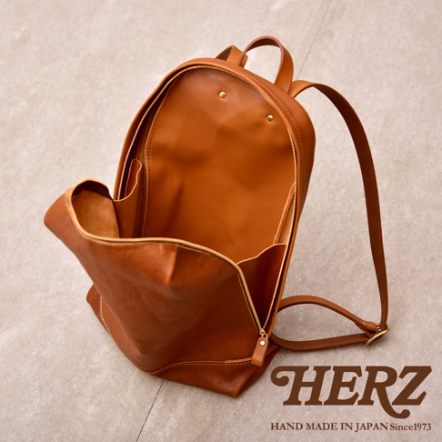 一張日本HERZ手工製作的Organ系列皮革後背包，以淺棕色義大利Eufrate皮革製成。畫面呈現包包開啟後的俯視角度，展現柔軟皮革的褶皺質感、簡約的內層空間及金色拉鍊細節。右下角標示HERZ品牌名稱與Hand Made in Japan字樣。