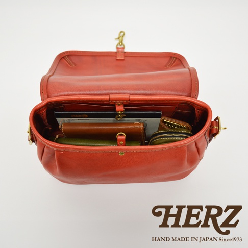 打開的橘紅色皮革復古翻蓋郵差包，內有筆記本、錢包、零錢包等多樣物品，包身線條圓潤，採用金屬扣環和縫線工藝。