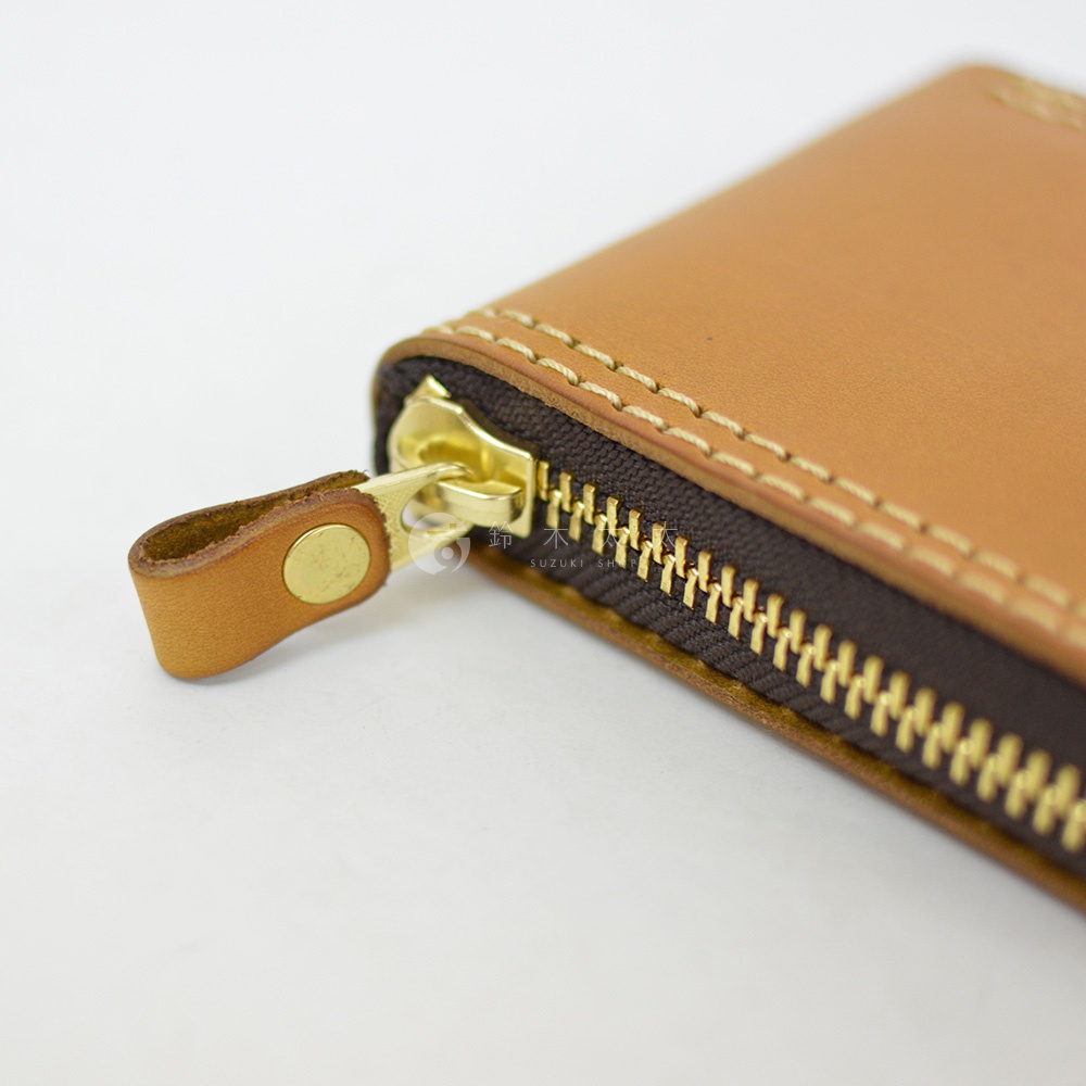 這是一張近距離拍攝的特寫照，焦點落在日本HERZ職人手作皮革短夾的金色拉鍊頭與深棕色布料拉鍊帶上。皮革表面呈現溫潤的淺棕色澤，邊緣以細緻的縫線進行加固，整體工藝質感細膩，呈現出高品質手工製品的細節特徵。
