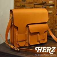 職人手作皮革單肩包，採用厚實的棕褐色皮革製成，包身有兩個前置翻蓋口袋，並以白色縫線裝飾，配有可調節的皮革肩帶。