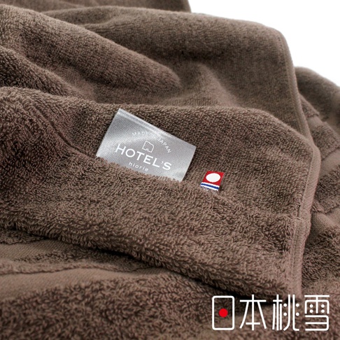 一條深棕色、柔軟的毛巾被，表面有細緻的絨毛紋理。毛巾被上縫有一塊銀色標籤，上面印有「HOTEL'S」字樣和「MADE IN JAPAN」的字樣，以及一個小巧的紅色、白色和藍色組成的徽標。