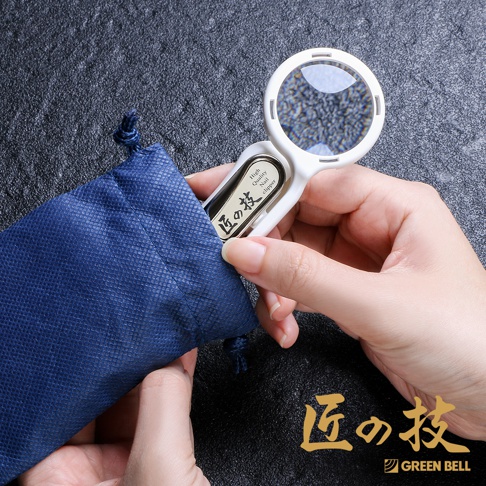 一隻手拿著白色不鏽鋼附放大鏡指甲剪，置於深藍色束口袋旁。指甲剪的刀刃部位嵌入袋口，放大鏡清晰可見。