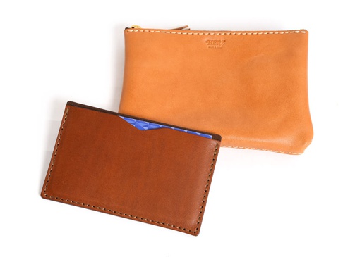 雙層皮革收納組，包含一個淺棕色卡夾，夾內有一張藍色幾何圖案卡片，以及一個較大的橘棕色拉鍊零錢包，兩者皆有白色縫線裝飾，材質皆為皮革。