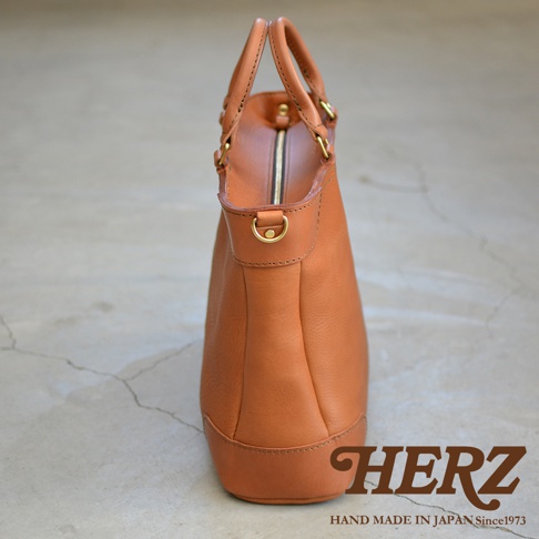 職人手作皮革Organ兩用商務托特包，呈現溫潤的棕褐色，包身由堅韌的皮革製成，縫線清晰。頂部設有提把和拉鍊開合，側邊有金屬D型環，結構紮實，適合商務場合。