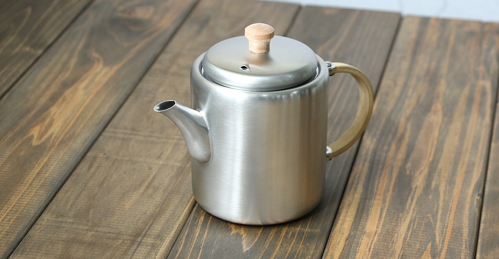 霧面不鏽鋼直筒茶壺，配有木質側邊把手與壺蓋拉環，簡約造型，放置於木紋桌面。