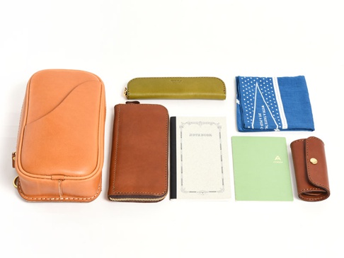 棕褐色皮革直立型斜背包，搭配綠色皮革筆套、棕褐色皮革長夾、藍色印花手帕、白色筆記本和綠色筆記本。