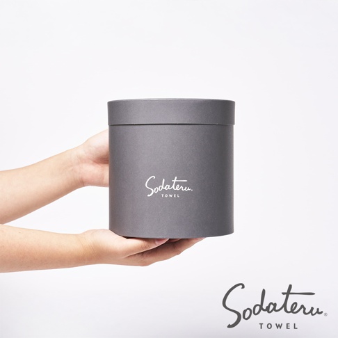 雙手捧著一個灰色圓筒形禮盒，盒身印有白色「Sodateru TOWEL」字樣。