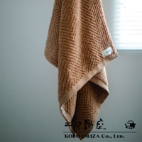 一條棕褐色人字紋有機棉浴巾，觸感柔軟，邊緣有米色滾邊，角落處縫有白色品牌標籤。