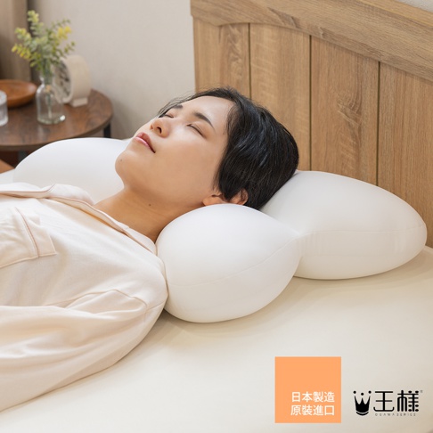 一位女性正平躺在床上，頭部舒適地枕在一個白色的王樣夢枕上，枕頭特殊的凹槽與弧形設計完整包覆支撐頭頸部，右下方標示著日本製造原裝進口以及王樣品牌字樣與皇冠標誌，畫面呈現出居家休憩的氛圍。