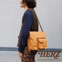 淺棕色皮革製成的厚實單肩包，包身有兩個翻蓋口袋，縫線明顯，由日本品牌 HERZ 製造。