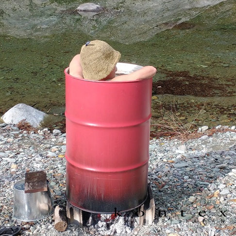 一個戴著淺棕色口袋桑拿帽的人，坐在紅色的圓筒狀物體中，置於河邊的鵝卵石地上，下方有火苗。