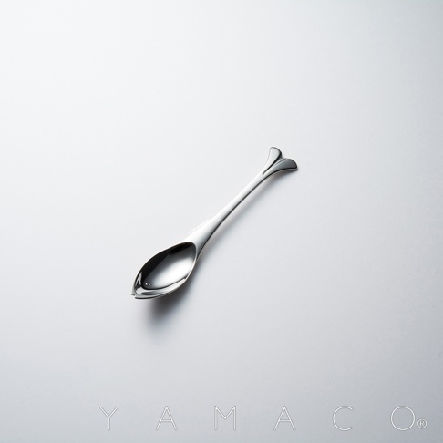 YAMACO 標誌旁有一把不鏽鋼魚形點心匙，勺頭設計成魚形，尾部呈魚鰭狀，光滑的銀色金屬反射著光線，放置在純白色背景上。