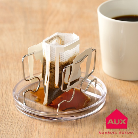 銀色金屬支架固定著滴漏式咖啡濾袋，濾袋內裝有咖啡粉，下方是透明圓形塑膠滴盤，盤中盛有滴漏的咖啡液，背景為木質桌面，右側有一白色咖啡杯。