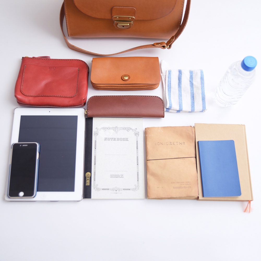 一組裝滿物品的日本HERZ皮質包，包含紅色和棕色皮夾、平板電腦、手機、筆記本、毛巾和水瓶。