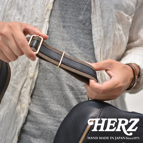 職人手作皮革多功能收納斜背包，黑色皮革包身，搭配咖啡色帆布與黑色皮革拼接的肩帶，肩帶上飾有金色金屬調節環。