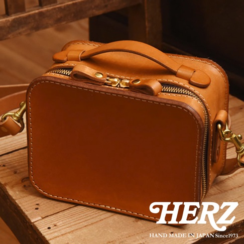 HERZ 品牌手工製作的棕色皮革小方包，配有金色拉鍊和掛鉤，展現職人精神。