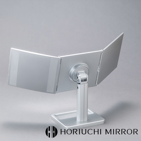 銀色金屬材質的桌上型三折化妝鏡，鏡面可360度旋轉調整，左側鏡面有碳纖維紋理裝飾，置於方形底座上。