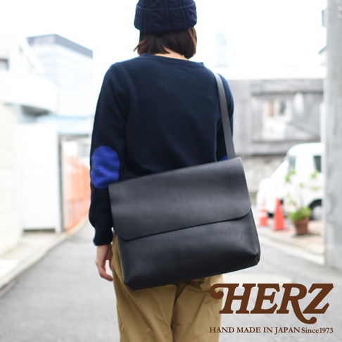 黑色皮革經典郵差包，配有可調節肩帶，展示於穿著海軍藍毛衣和卡其色褲子的模特身上。