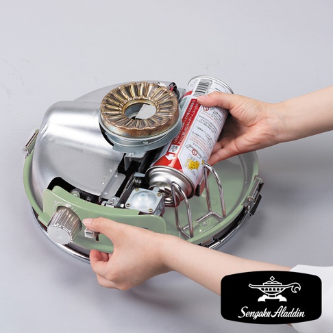 雙手正在將卡式瓦斯罐裝入淺綠色金屬材質的瓦斯暖爐主體。暖爐頂部有銀色金屬燃燒器，側邊有一個銀色旋鈕。