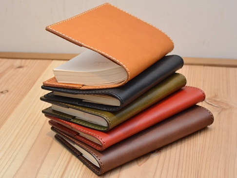 堆疊的職人手作皮革厚革書套，從上至下顏色依序為：淺棕色、黑色、墨綠色、磚紅色、深棕色。書套由皮革製成，邊緣縫線清晰，頂部一個書套內含一本白色紙張的筆記本。
