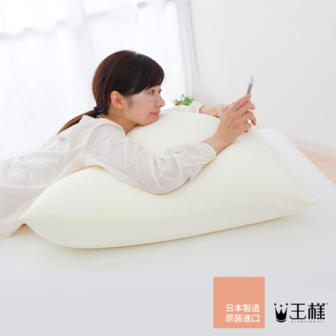 一位女性側臥在淡黃色的巨大枕頭上，枕頭材質看起來柔軟，女性穿著白色長袖上衣，手持手機。