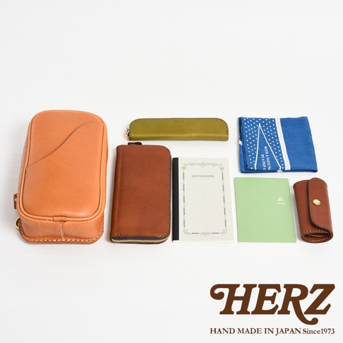 日本HERZ品牌手工皮革製品，包含一個棕色斜背包、一個橘色長夾、一個綠色筆袋、一本筆記本、一個綠色筆記本和一個棕色鑰匙包，下方有HERZ品牌標誌和「HAND MADE IN JAPAN Since1973」字樣。