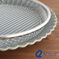 銀色金屬網格濾網，底部置於藍灰色波浪邊緣的陶瓷盤上，背景為木質桌面。