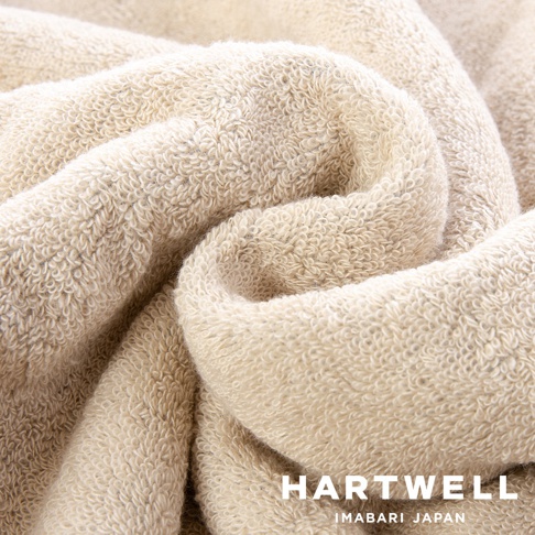 這是一張特寫鏡頭，展示了 HARTWELL 今治毛巾的細膩紋理，米色調的織物呈現出柔軟且蓬鬆的圈絨質感，透過優雅的捲曲摺痕突顯出高品質的布料結構，右下角印有 HARTWELL IMABARI JAPAN 的標誌。