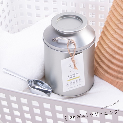 銀色牛奶罐造型洗衣粉罐，附有標籤及金屬量勺，置於白色洗衣籃中，背景為白色毛巾及木紋紋理。