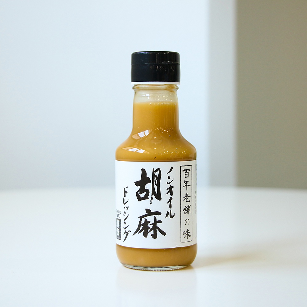 玻璃瓶裝無油芝麻沙拉醬，醬體呈現淺棕色，瓶身貼有白色標籤，上面印有黑色日文與中文書法字體，標示產品名稱與品牌資訊，瓶蓋為黑色。