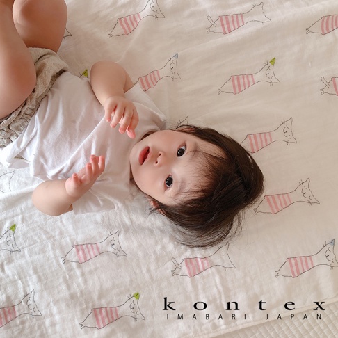 嬰兒躺在印有可愛貓咪圖案的白色二層紗嬰兒毯上，貓咪圖案為粉色條紋，部分貓咪頭部有綠色或藍色點綴。