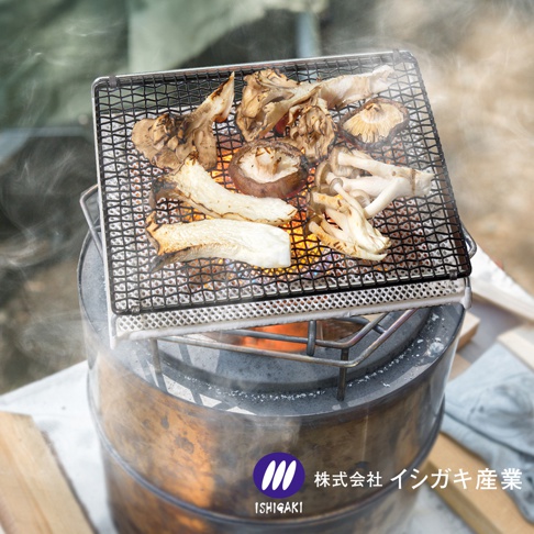 一個金屬質感的圓形露營瓦斯爐上放置著方形陶瓷烤網，網架上擺放著數種正在燒烤的各式菇類，爐身印有石垣產業的企業標誌，整體呈現出戶外野炊的情境。