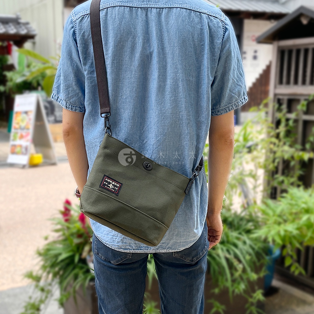 橄欖綠純綿帆布迷你側背包，附有深棕色可調式斜背帶，包身正面有方形品牌布標和一個圓形鈕扣。