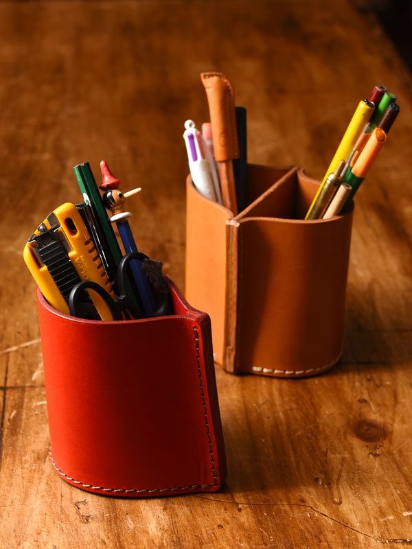 兩個職人手作皮革筆筒，一個為亮紅色，另一個為棕褐色。紅色筆筒內裝有美工刀、剪刀、鉛筆及其他文具；棕褐色筆筒則有分格，內放有多支不同顏色的筆。