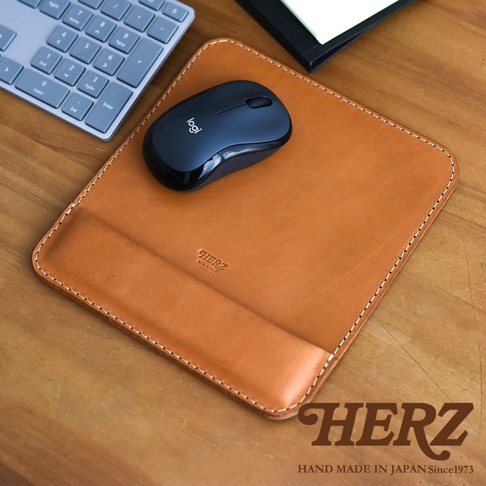 駝色皮革滑鼠墊，帶有手腕支撐設計，邊緣以白色縫線處理，上面放置了一個黑色無線滑鼠。