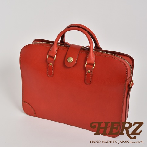 紅色皮革製成的手提公事包，有雙提把、金色金屬扣和拉鍊。包款設計簡潔，邊角處有皮革加固。