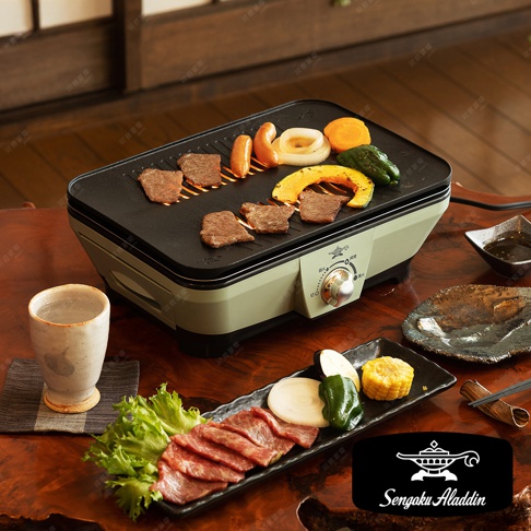 這是一台淺綠色與黑色搭配的迷你烤盤，烤盤上烤著牛肉、香腸、洋蔥、櫛瓜和南瓜。烤盤下方有一個銀色的旋鈕和標示，側面有產品名稱「Sengoku Aladdin」的標誌。