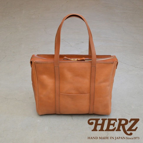 焦糖色皮革製成的肩背拉鍊托特包，包身有縫線細節與一個外層口袋，搭配同色系皮革提把，整體呈現簡約風格。