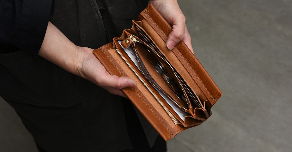 一位穿著深色衣物的人雙手捧著一個敞開的日本HERZ職人手作皮革長夾，該長夾呈現溫潤的棕色皮革質感，內部設有拉鍊零錢袋並裝有鈔票與卡片，整體設計簡約實用。