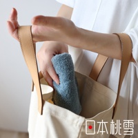 一隻手將一條淺藍色、柔軟的毛巾放入米白色帆布袋中，毛巾質地蓬鬆，袋子配有淺棕色皮革提把。