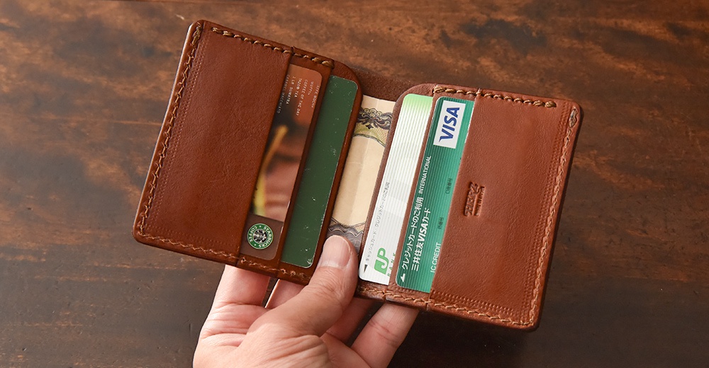 職人手作皮革二折式薄型卡片鈔票夾，展示卡夾內有信用卡、日圓鈔票及綠色卡片。