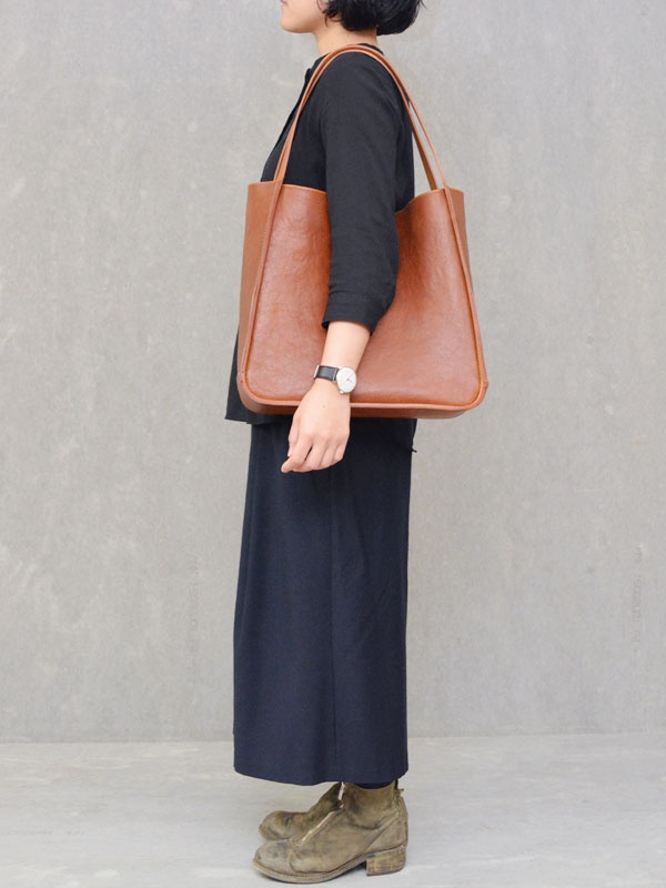 一位女士身穿黑色長袖上衣和深藍色長裙，左肩斜挎一個棕色方形皮革托特包，包身線條簡潔，配有細長的肩帶。