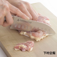 鉬釩不鏽鋼三德刀，刀刃上有橢圓形凹槽，正在切塊的粉色生雞肉，置於淺木色砧板上。