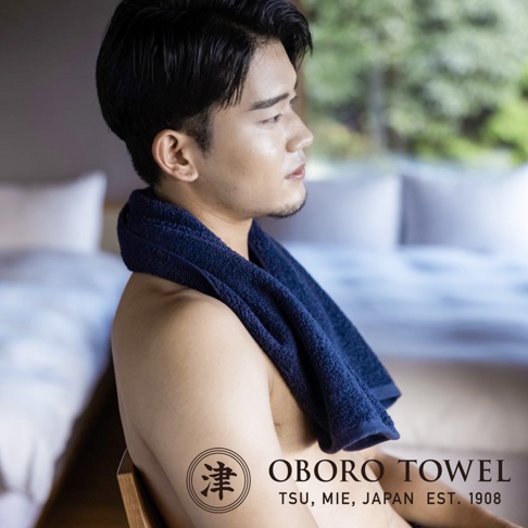 一名男子將深藍色毛巾搭在肩上，背景是模糊的臥室。毛巾材質看起來柔軟，並帶有細緻的紋理。