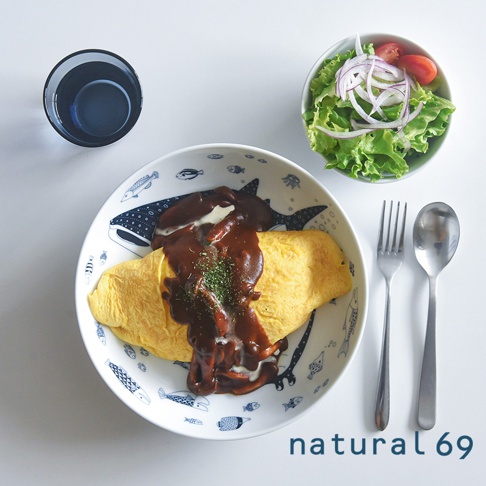 一個裝有蛋包飯的白色圓形深碗，碗內有藍色魚紋圖案，蛋包飯上淋有褐色醬汁和白色醬汁，並撒有綠色香料。碗旁有餐巾、叉子、湯匙和一杯深藍色飲料。上方還有一小碗沙拉，內有生菜、番茄和洋蔥。