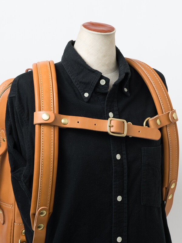 職人手作皮革背包用減壓胸前扣帶，採用天然棕色皮革製作，搭配黃銅色金屬扣環與铆釘，固定於黑色襯衫的背包肩帶上。