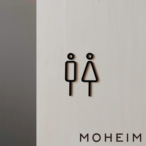 黑色簡約風格的男女廁所標示牌，由木質紋理的淺色背景襯托，上方有黑色線條勾勒的男性與女性符號，下方印有品牌名稱「MOHEIM」。